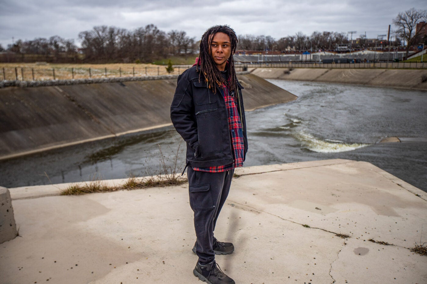 Marcell Simmons stands on a concrete ledge near the Flint River. He has his hands in his pockets and stares off camera left.