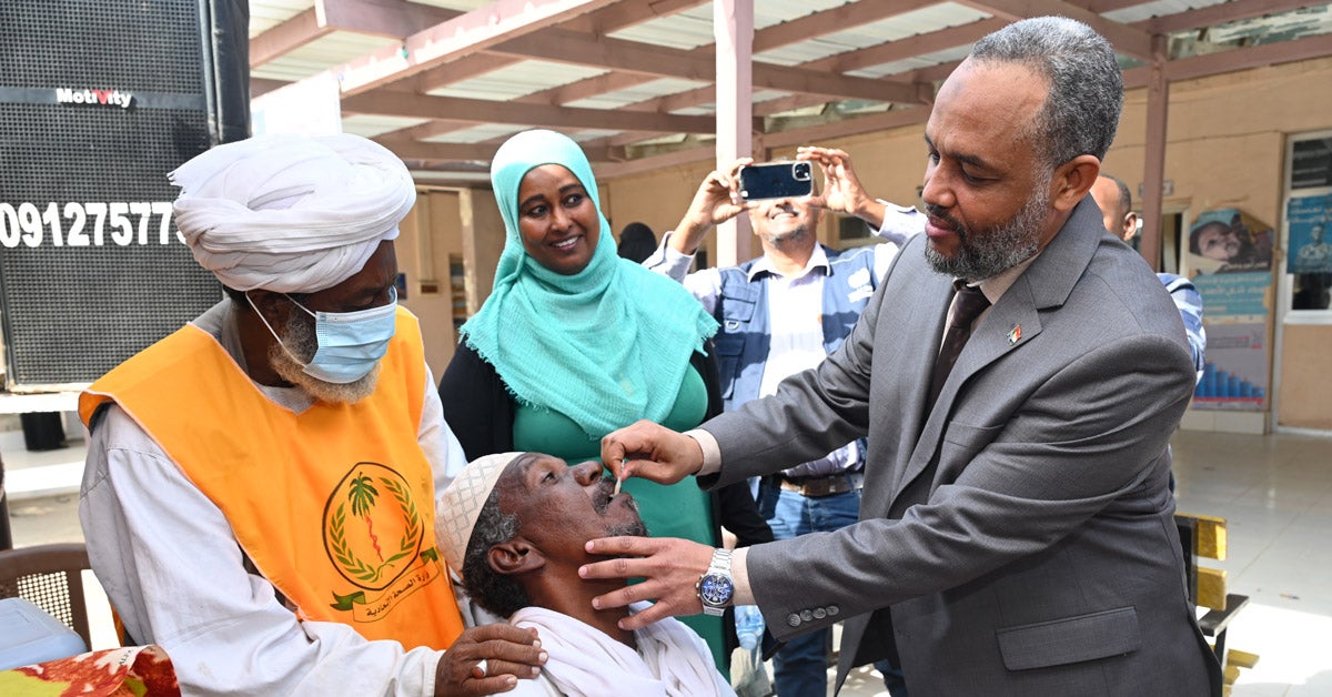 Heitham Mohammed Ibrahim Awadalla, Sudan’s minister of health, administers an oral vaccine to an adult man at a vaccine event. A healthcare worker puts his hands ont he patient’s shoulders. Two other people look on and smile, taking photos.