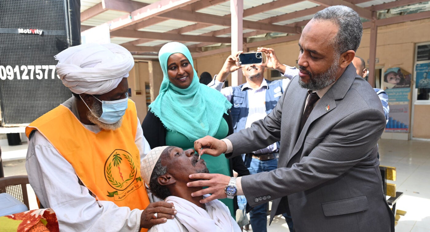 Heitham Mohammed Ibrahim Awadalla, Sudan’s minister of health, administers an oral vaccine to an adult man at a vaccine event. A healthcare worker puts his hands ont he patient’s shoulders. Two other people look on and smile, taking photos.