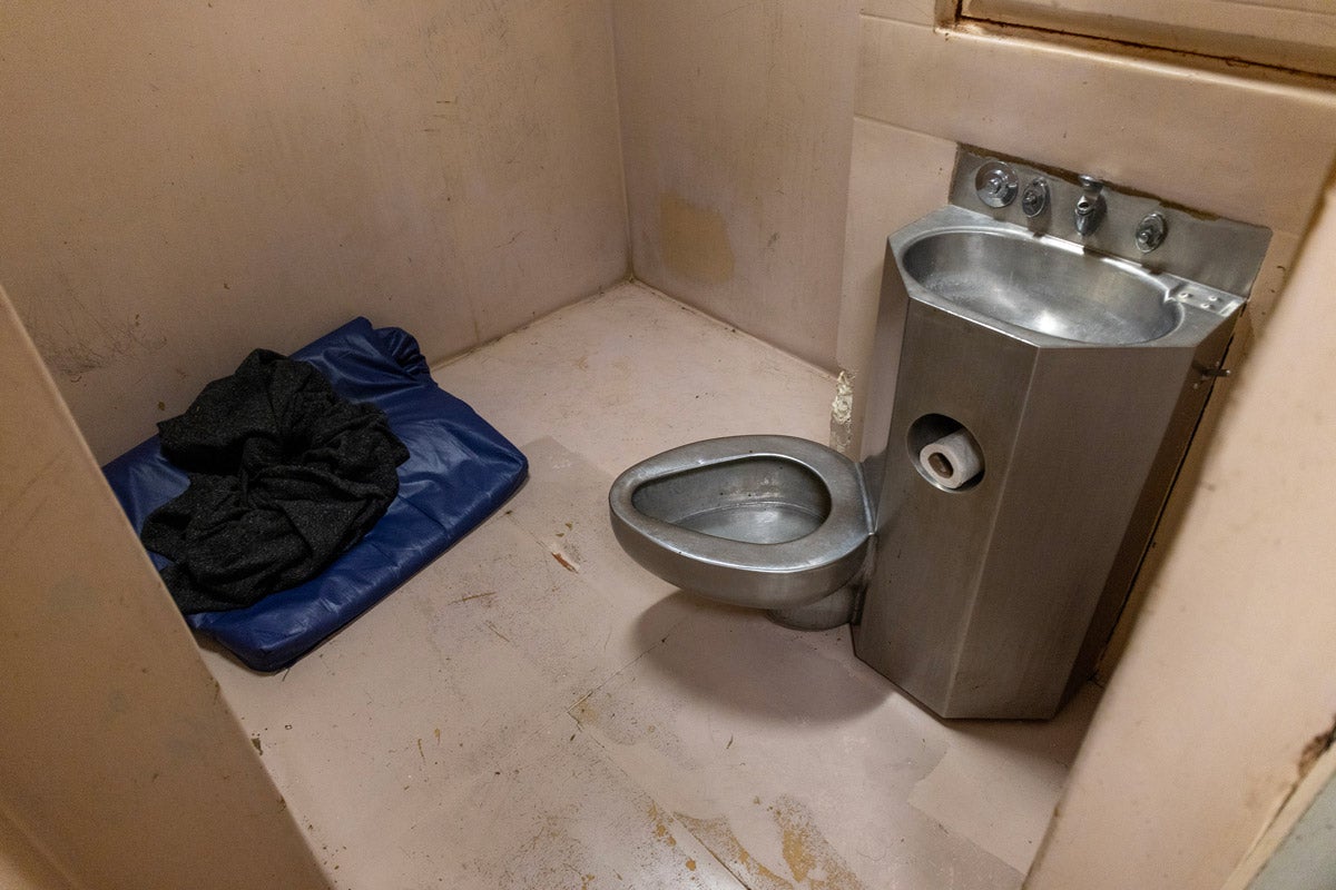 A padded jail cell with filthy, yellowing walls and a dirty floor has a metal sink and toilet attached to the wall. In the corner is a small blue sleeping bag and black blanket.