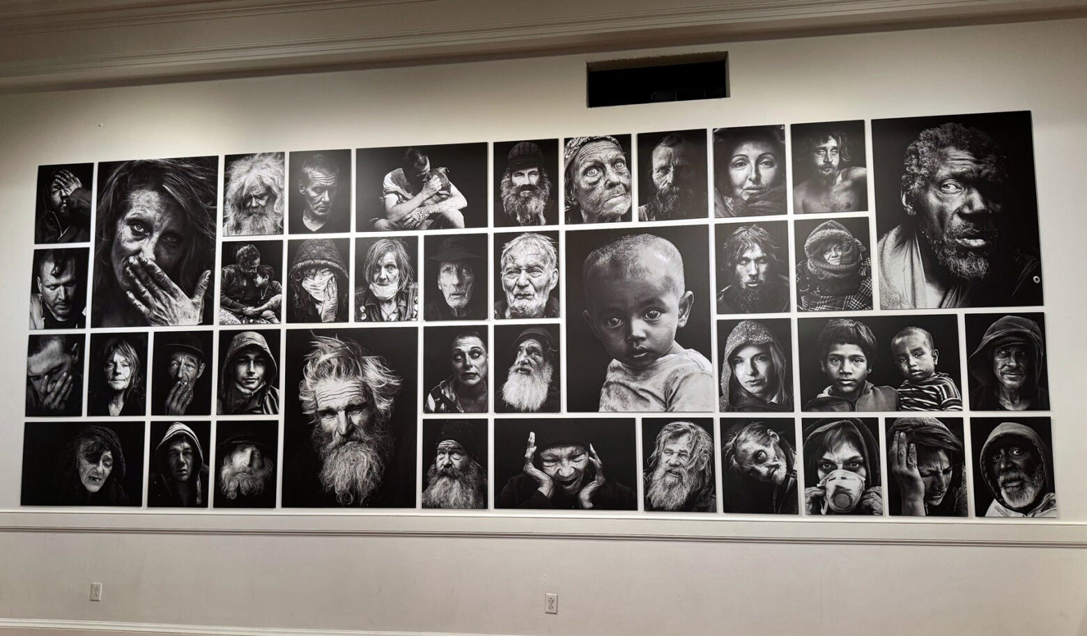 Forty black and white portraits hang on a gallery wall in a grid. The photos are of unhoused people in various expressions.