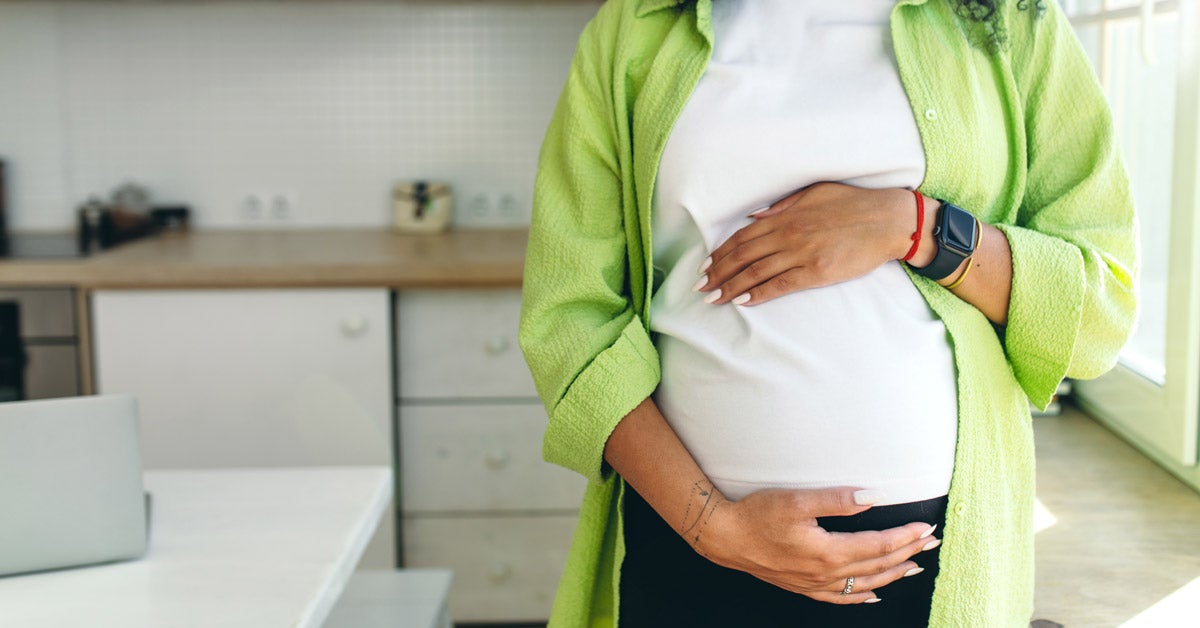 Pregnant Black woman with long curly hair stands in a kitchen and looks out a window. Her hands rest on her belly.