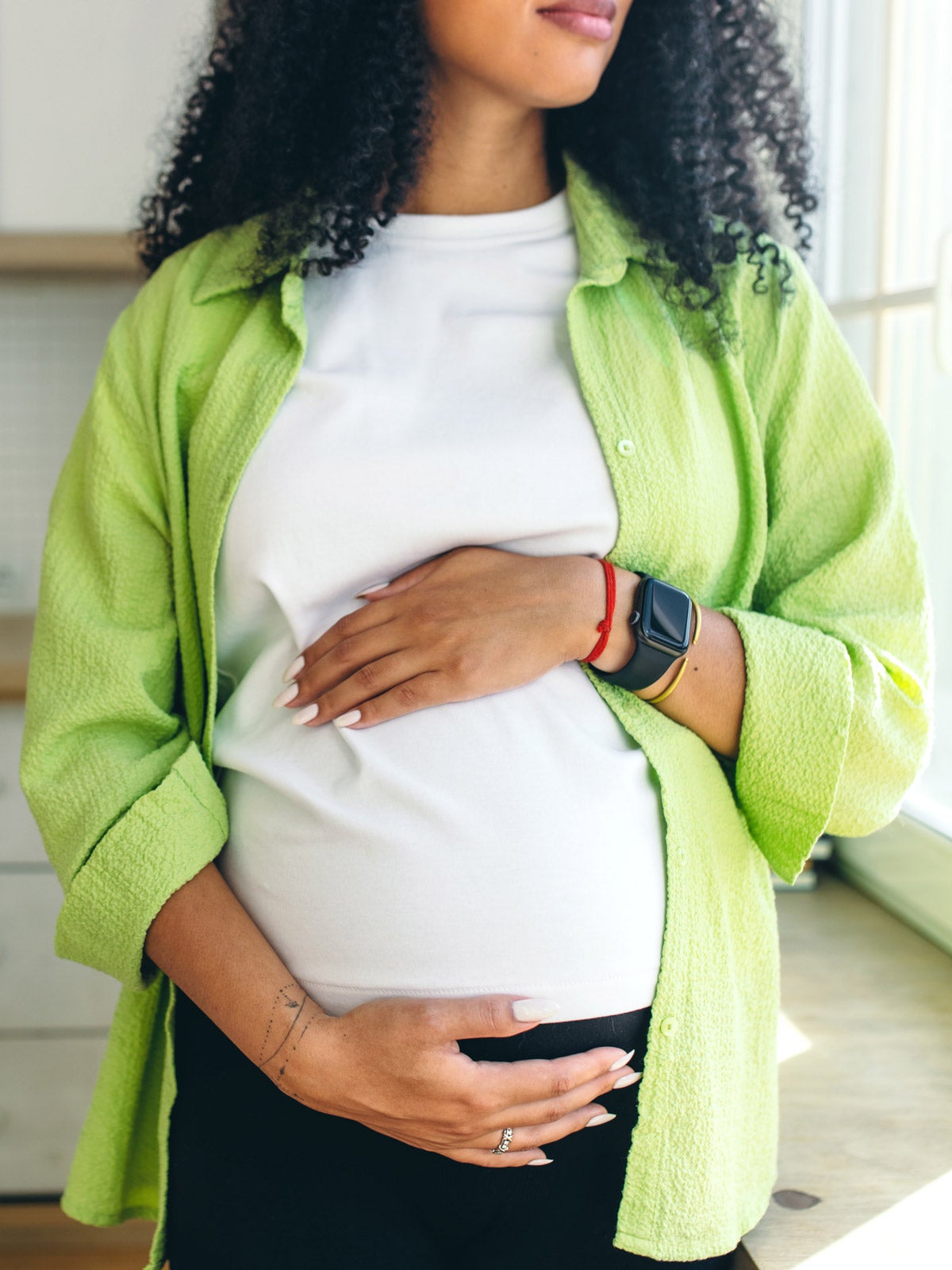 Pregnant Black woman with long curly hair stands in a kitchen and looks out a window. Her hands rest on her belly.