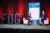 Michael Fitzgerald and Catalina Lopez-Correa in conversation on a conference stage. they sit in white chairs and red curtains hang behind them. There is also a banner with sponsors and the letters "FOG" which stand for Festival of Genomics on the stage.