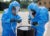 Two people in blue hazmat suits stand over a black bin in a parking lot.