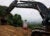 Tom Bussolett stands with his back to the camera near a cleared dirt patch in a forest. A black earth digger rests nearby.