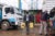 Mohammed Suleiman carries two yellow jugs through a busy street in Makina Kenya. He wears a backwards ball cap, jacket, white shirt and pants.