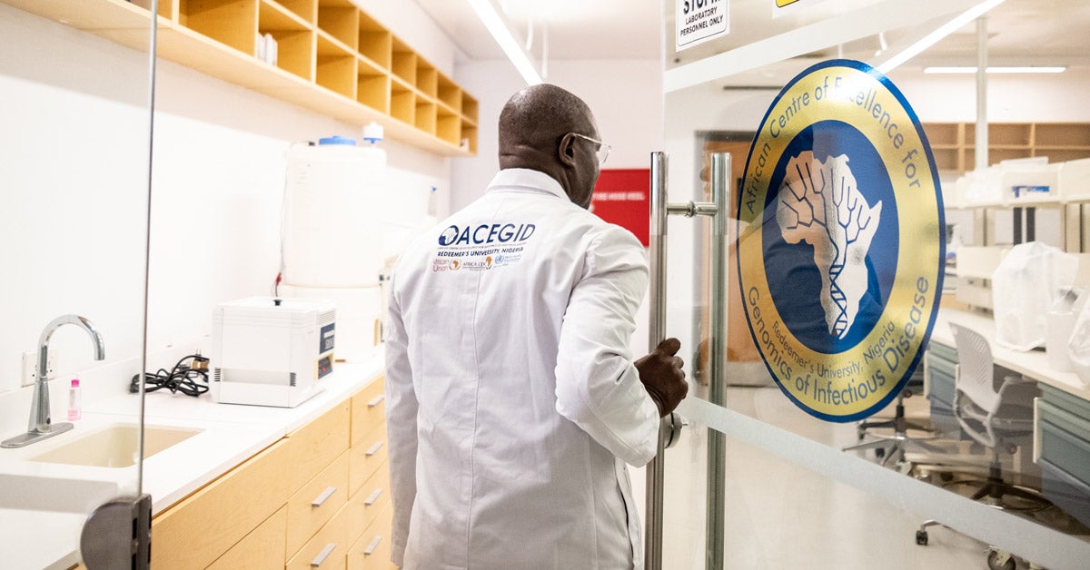 Christian Happi wearing an embroidered white lab-coat enters a dry lab through a set of clear double-doors at ACEGID.