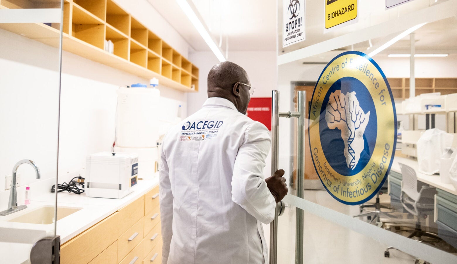 Christian Happi wearing an embroidered white lab-coat enters a dry lab through a set of clear double-doors at ACEGID.