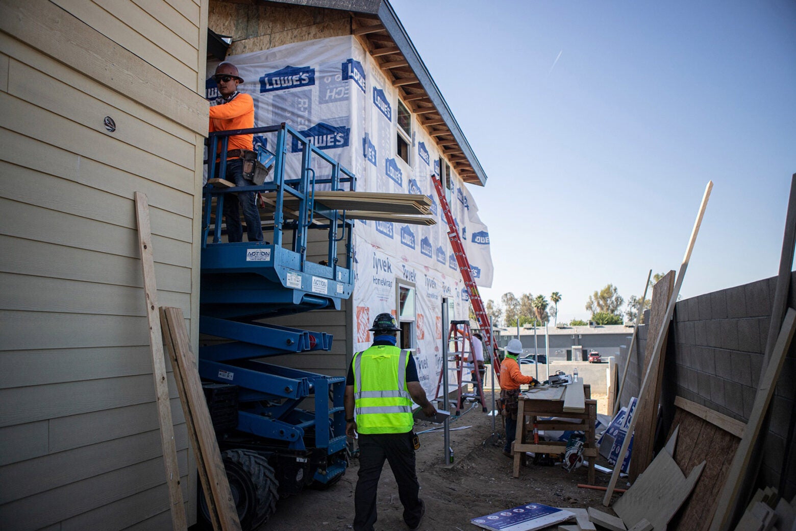 Construction workers dressed in bright green and orange outfits work on an apartment building. The building is clearly unfinished and covered in a plastic sheet.