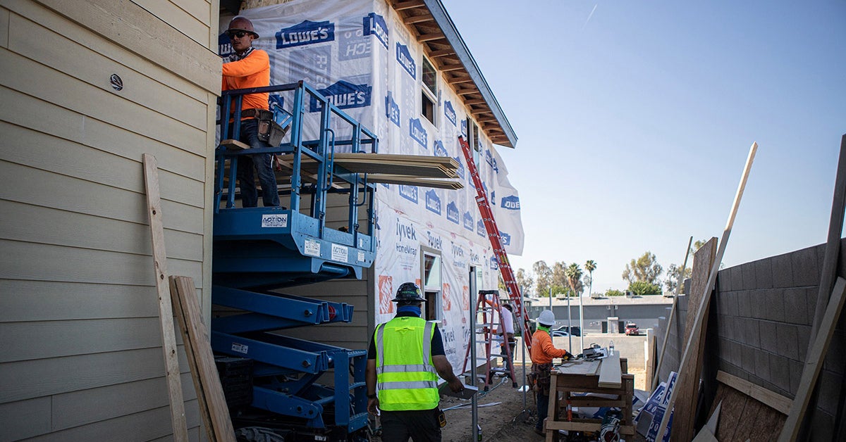 Construction workers dressed in bright green and orange outfits work on an apartment building. The building is clearly unfinished and covered in a plastic sheet.