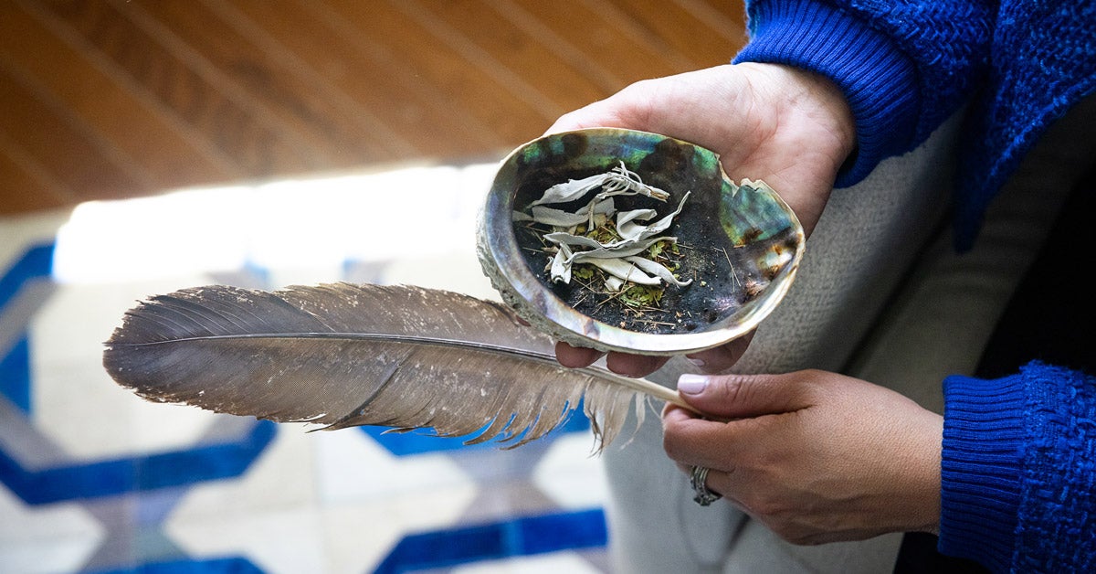 Two hands hold a small ceramic dish with dried flora and a bird feather. The figure is wearing a bright blue sweater.