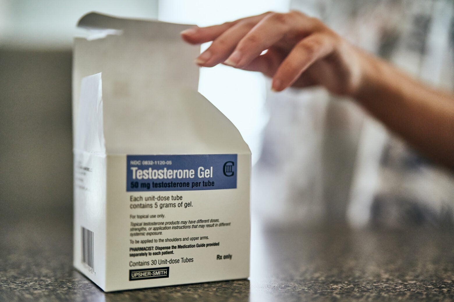 A box of open testosterone gel on a granite counter. A hand touches the box top.