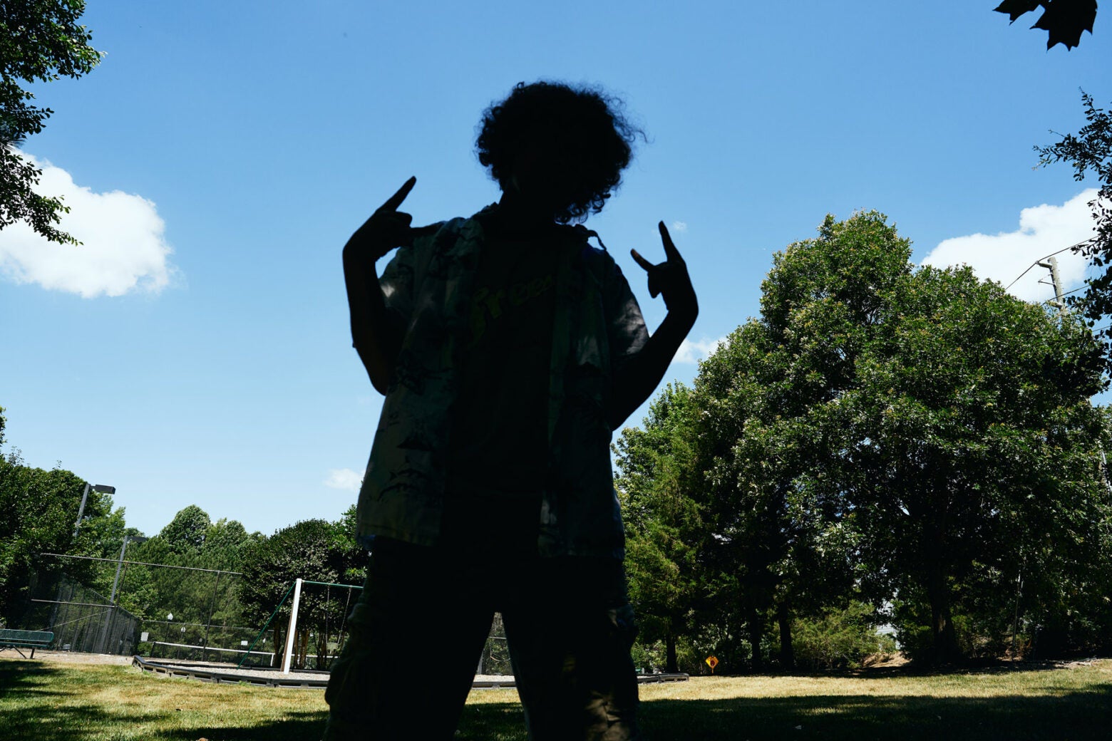 A teen stands in the middle of a grassy rec field and makes the "rock on" signs with his hands. His body is in silhouette.