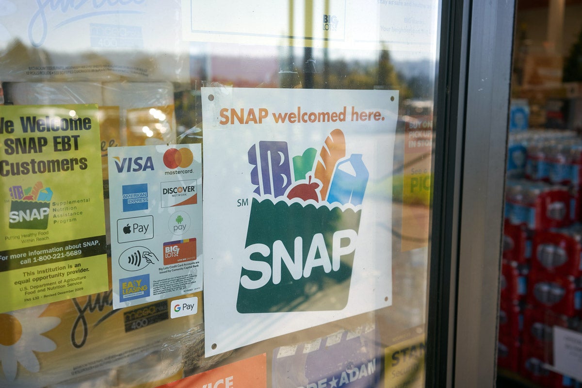 A white “SNAP welcomed here" sign on a store window. On it’s left is a credit card accepted sign with logos, and a yellow “We welcome snap EBT Customers” flyer.