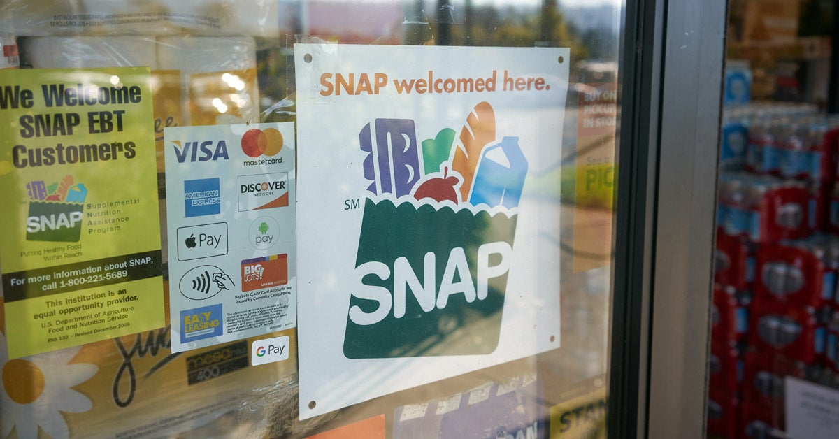 A white “SNAP welcomed here" sign on a store window. On it’s left is a credit card accepted sign with logos, and a yellow “We welcome snap EBT Customers” flyer.