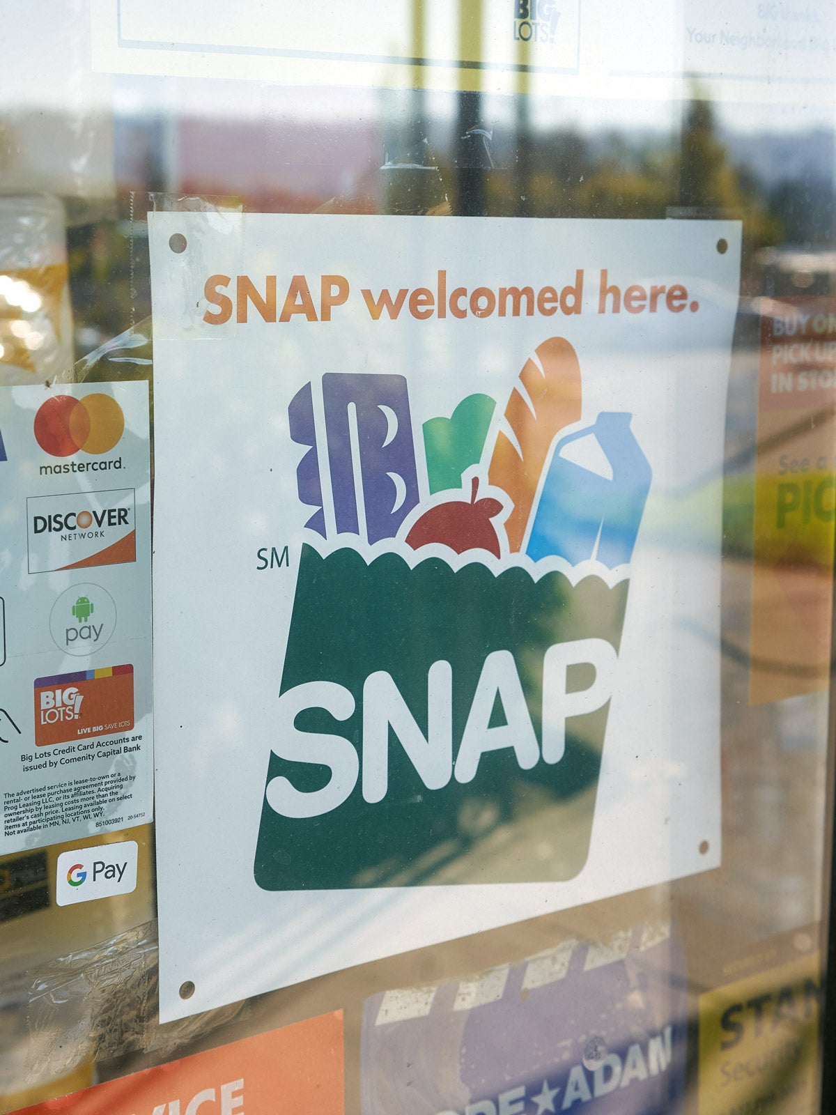 A white “SNAP welcomed here" sign on a store window. On it’s left is a credit card accepted sign with logos, and a yellow “We welcome snap EBT Customers” flyer.