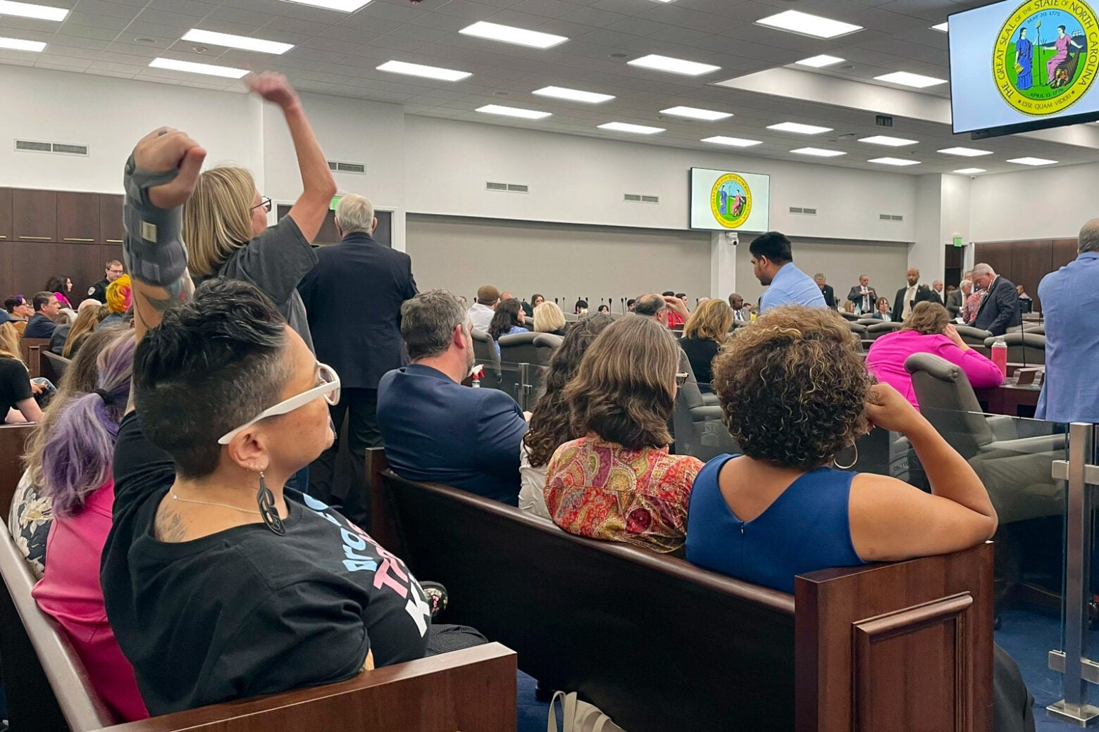 A group of women and men sit in a busy government meeting room in North Carolina. Some stand with arms raised.