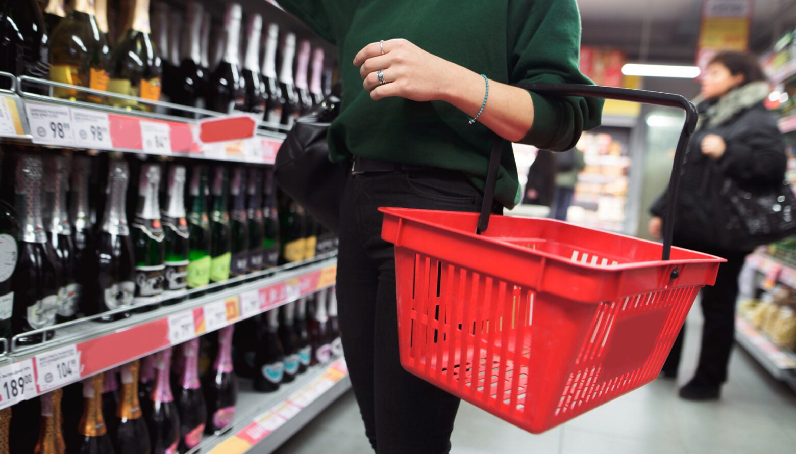 A woman in a green sweater with a red shopping basket chooses alcohol in a supermarket.