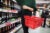 A woman in a green sweater with a red shopping basket chooses alcohol in a supermarket.