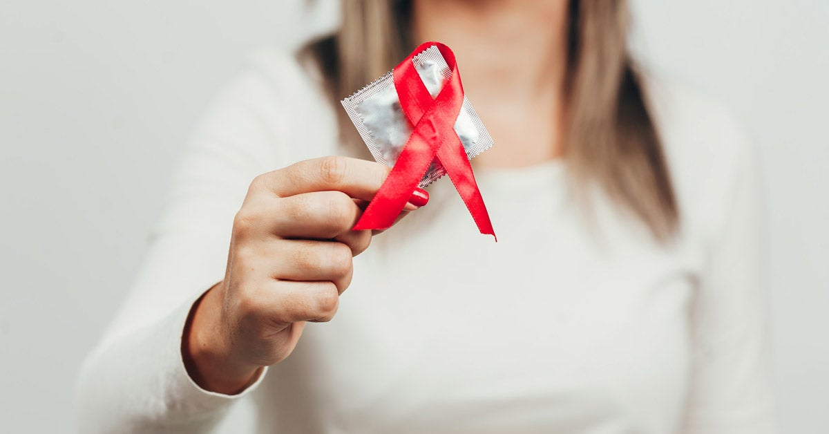 A woman, face out of view and body blurred, holds a condom with a red AIDS ribbon hanging off the corner.