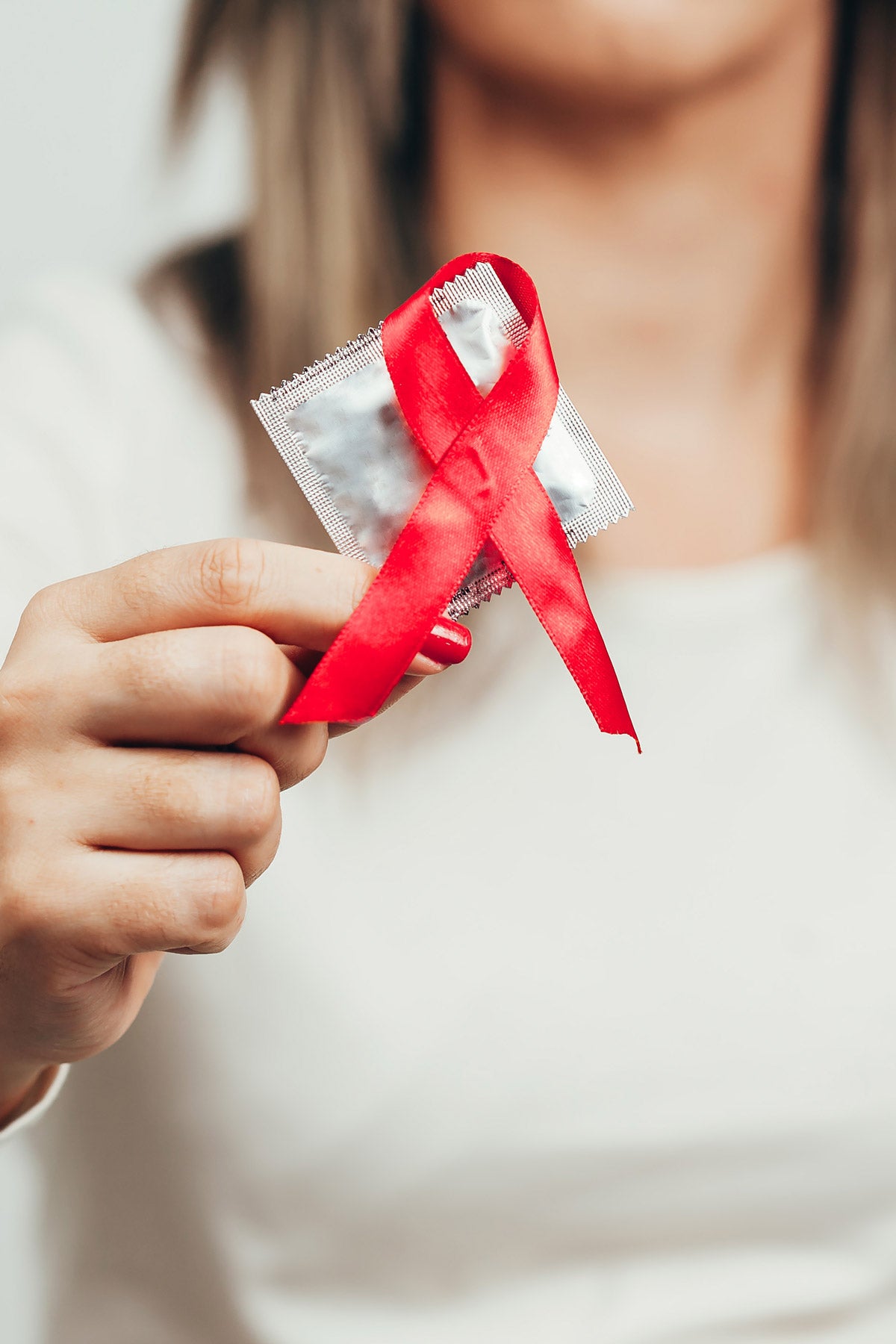 A woman, face out of view and body blurred, holds a condom with a red AIDS ribbon hanging off the corner.