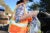 A woman in an orange reflective vest, baseball cap and mask, carries a bag of green apples to a black pick-up truck.