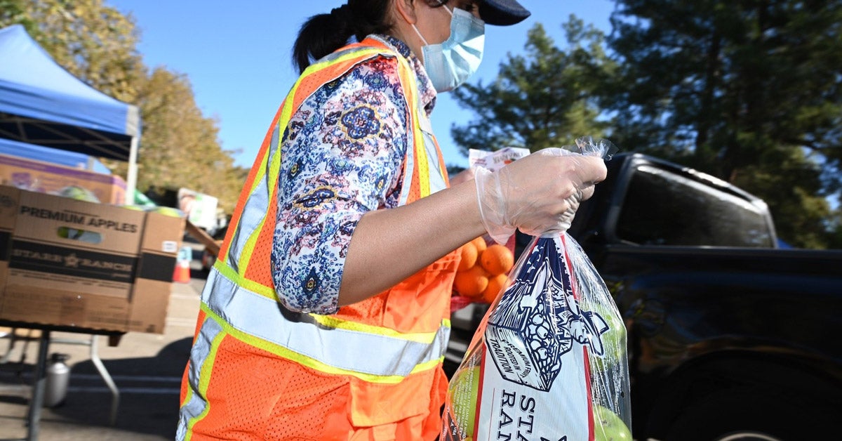 A woman in an orange reflective vest, baseball cap and mask, carries a bag of green apples to a black pick-up truck.