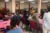 Lorena Estrada-Martinez leads and listens to a group of citizens. She stands while others sit in red chairs at pink table-clothed long tables.