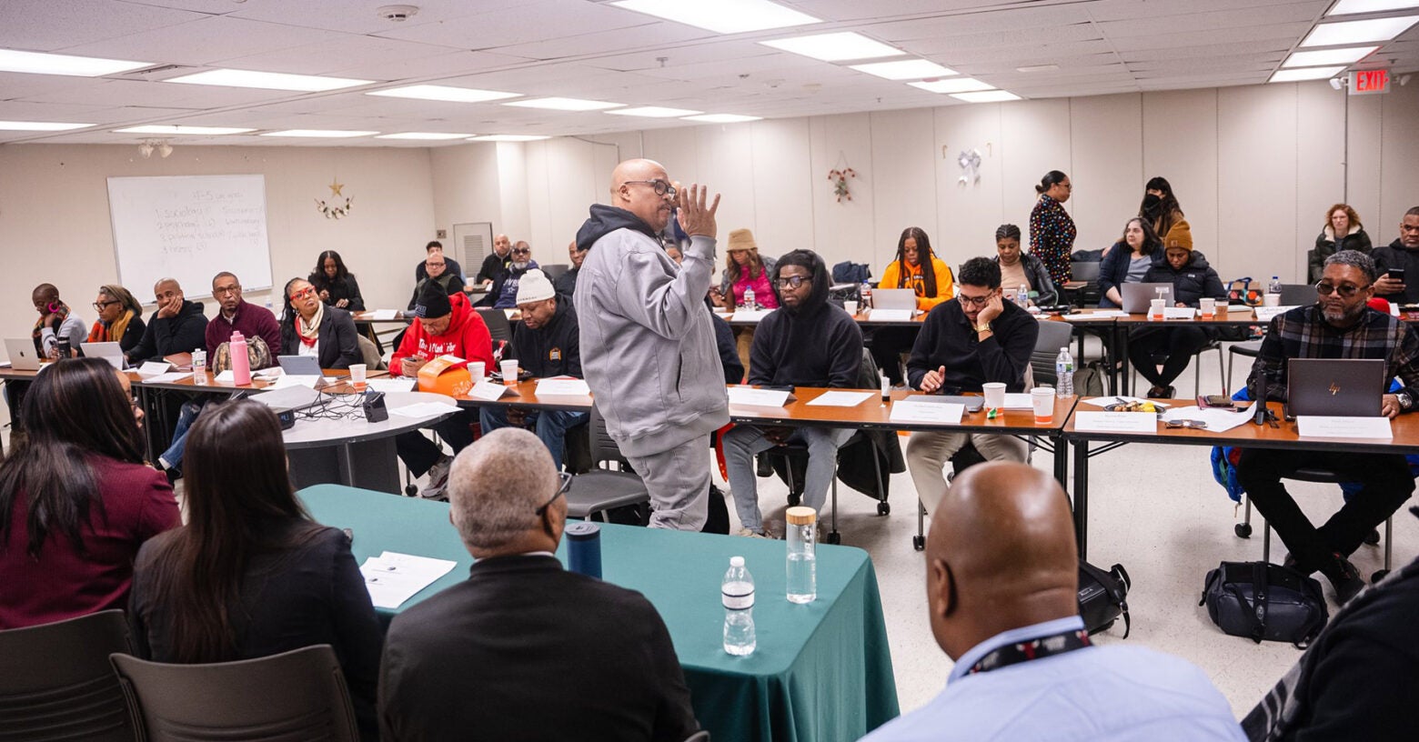 Marcus MacAllister speaks to a classroom full of adult students and learners. Students sit at long tables arranged in rows.