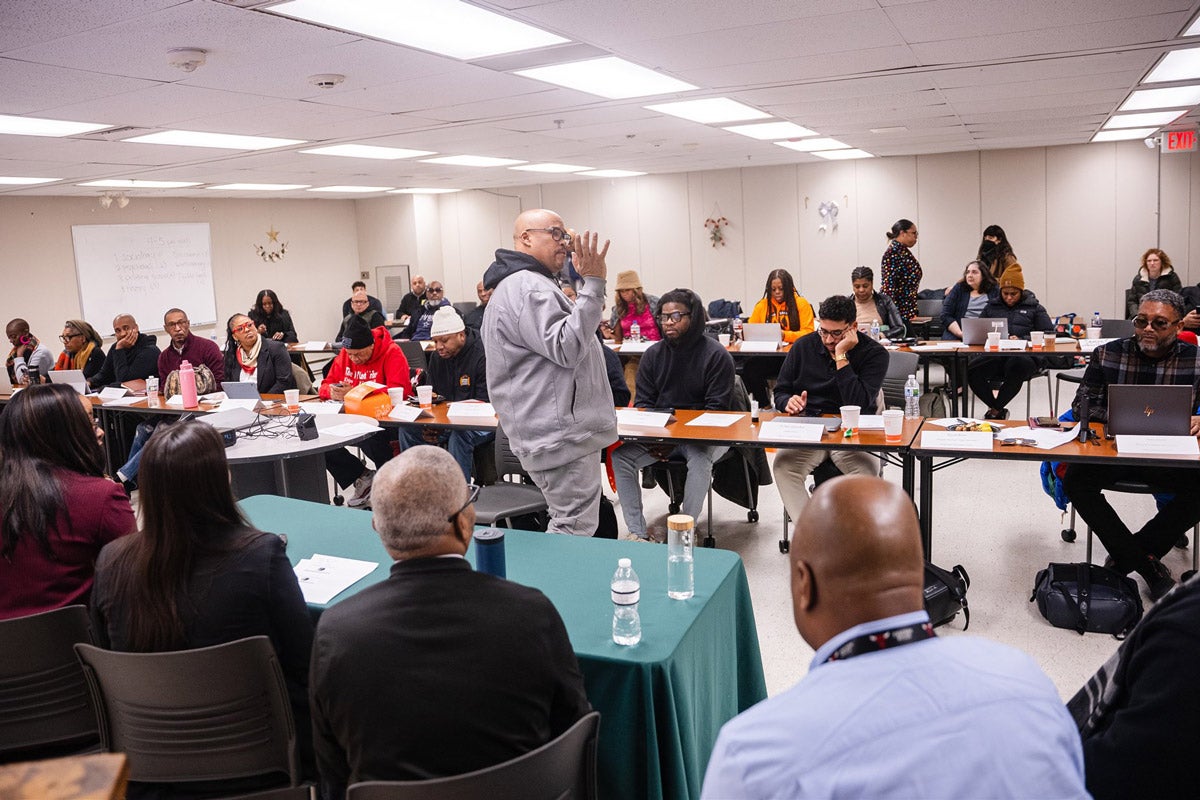 A trainer specializing in community violence intervention speaks to a classroom full of professionals working in the field of violence intervention. Students sit at long tables arranged in rows.