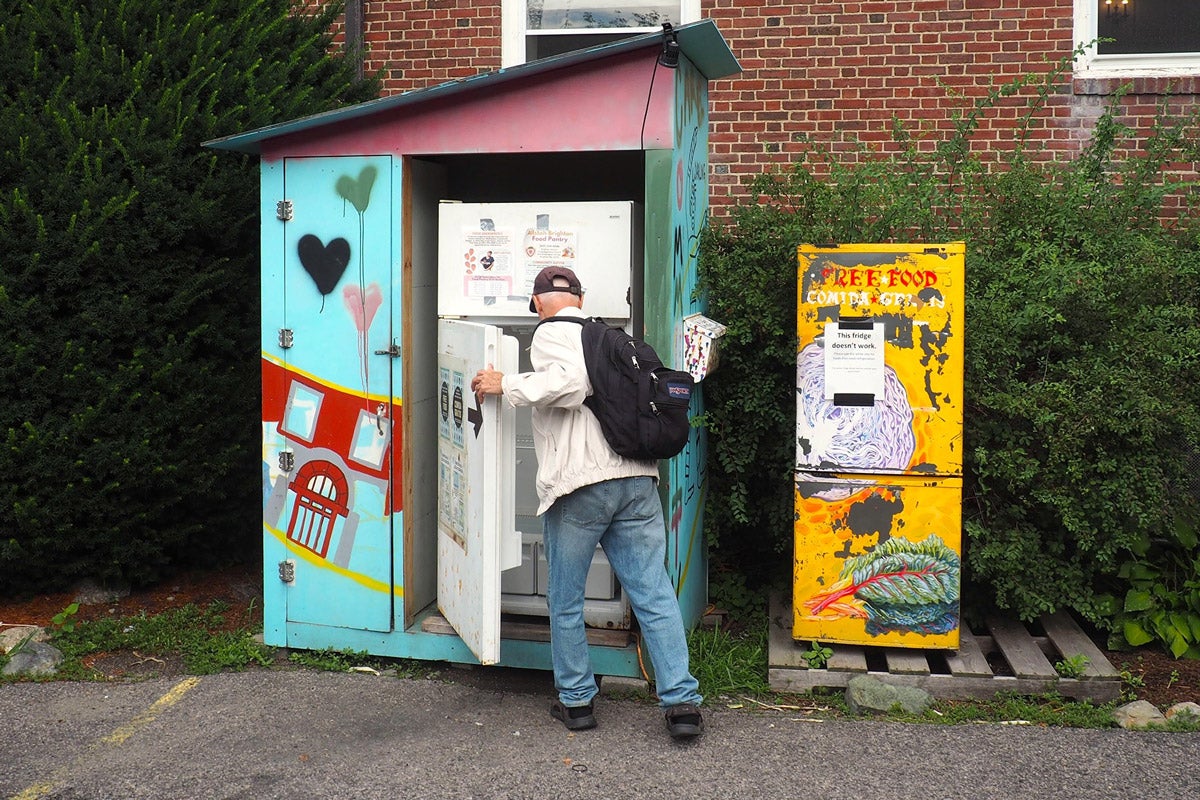 An older man opens a white refrigerator sitting outdoors next to some bushes in the city. It is housed in a blue shed decorated with colorful art.