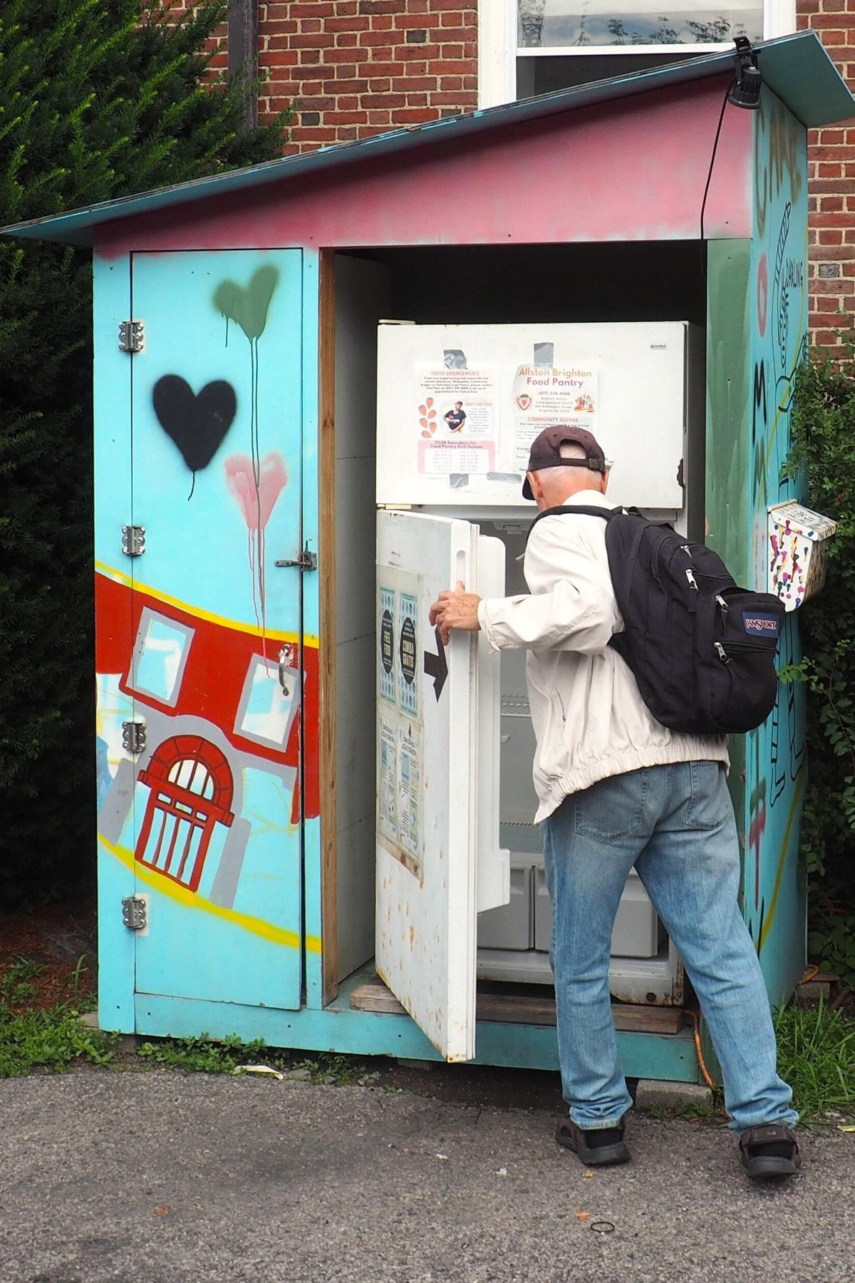 An older man opens a white refrigerator sitting outdoors next to some bushes in the city. It is housed in a blue shed decorated with colorful art.