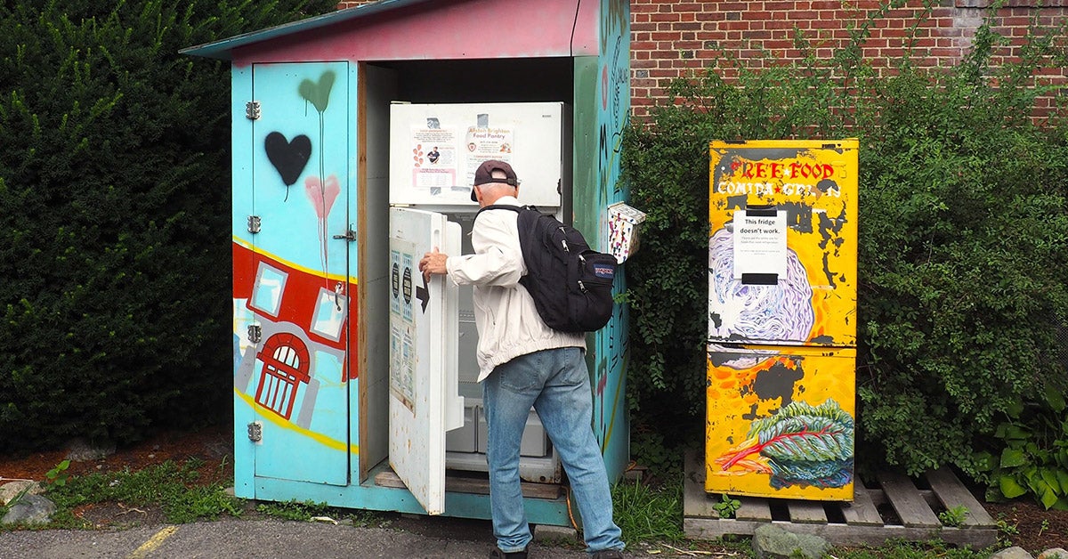 An older man opens a white refrigerator sitting outdoors next to some bushes in the city. It is housed in a blue shed decorated with colorful art.