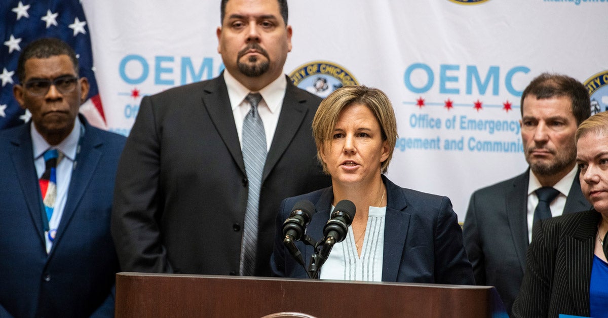 Jennifer Layden speaks into two microphones at podium at a city of Chicago press conference. Four other officials stand behind her.