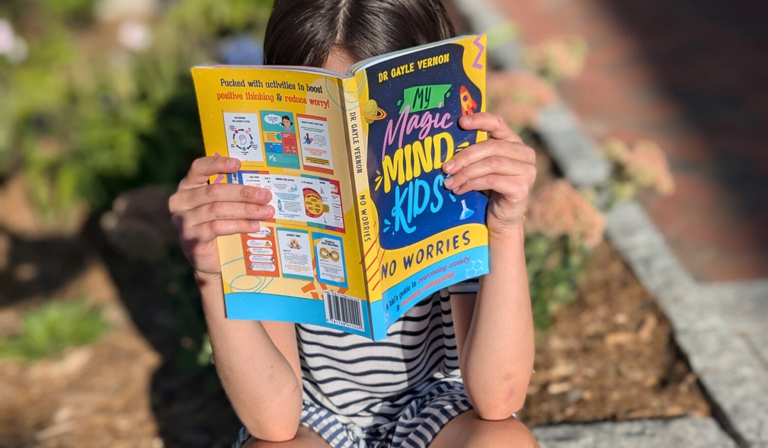 A girl holds sits outside and reads "My Magic Mind Kids: No Worries"