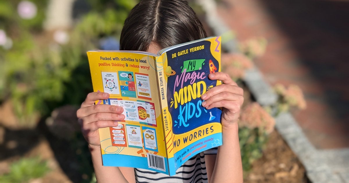 A girl holds sits outside and reads "My Magic Mind Kids: No Worries"