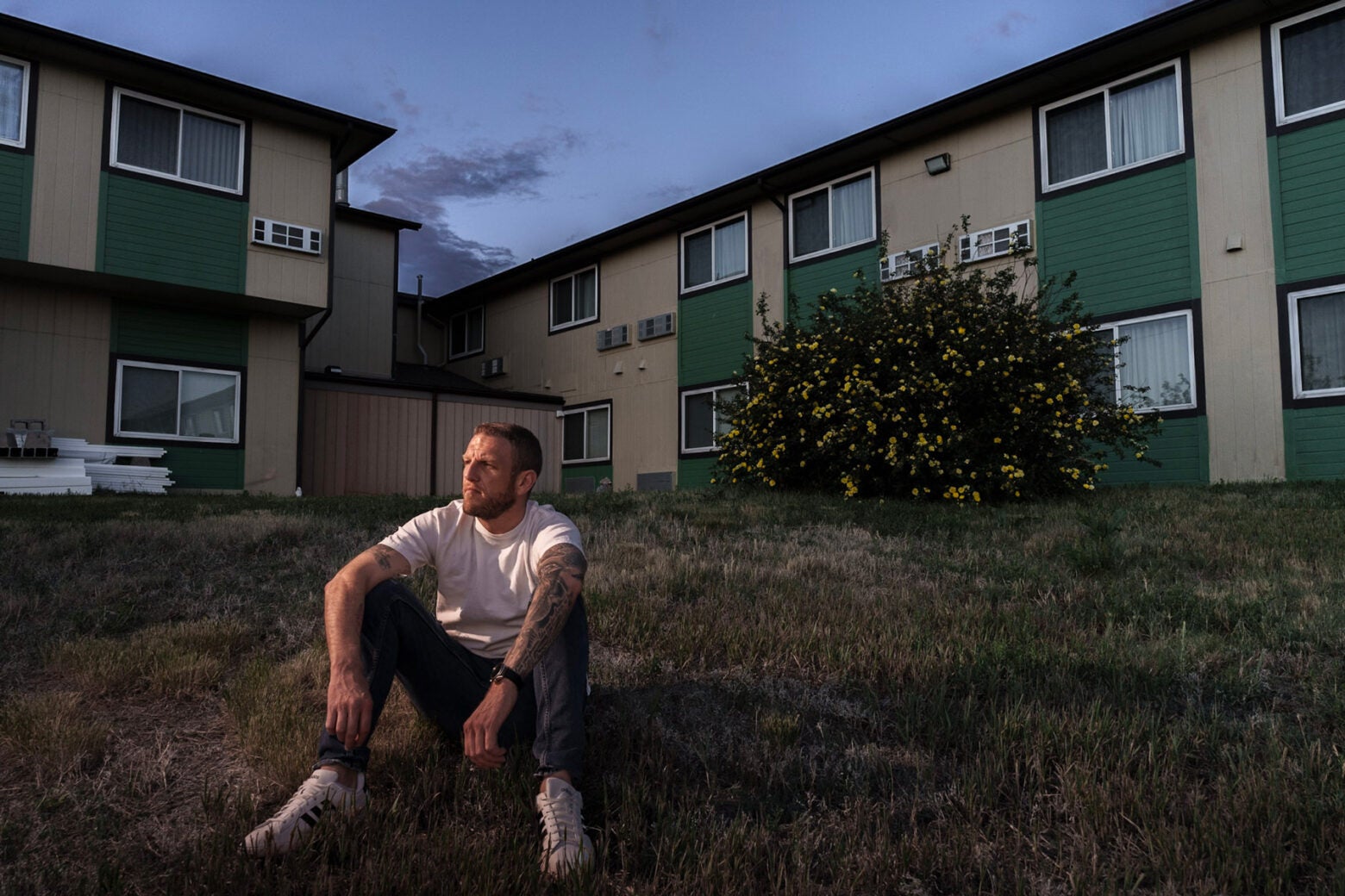 A middle-aged white man with tatoos on one arm, sits on a grassy hill outside a green and tan hotel.