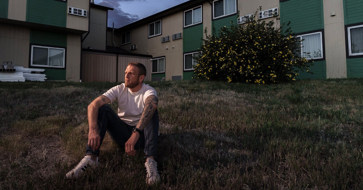 A middle-aged white man with tatoos on one arm, sits on a grassy hill outside a green and tan hotel.