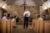 A middle-aged man in a white shirt and tie stands in the middle of a church aisle and talks to four family members in pews. A cross and stone facade are behind him.