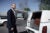 A middle-aged white man wearing a suite and tie looks up at the sky. He stands besides a white hearse with the door open revealing a gray interior and a brown casket.