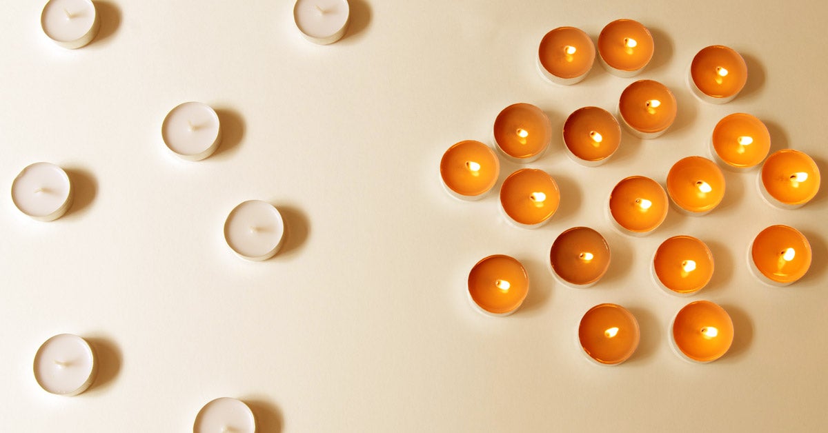 Seven unlit, white votive candles are separated on one side of surface. On the other, a cluster of lit, orange votive candles huddle together.