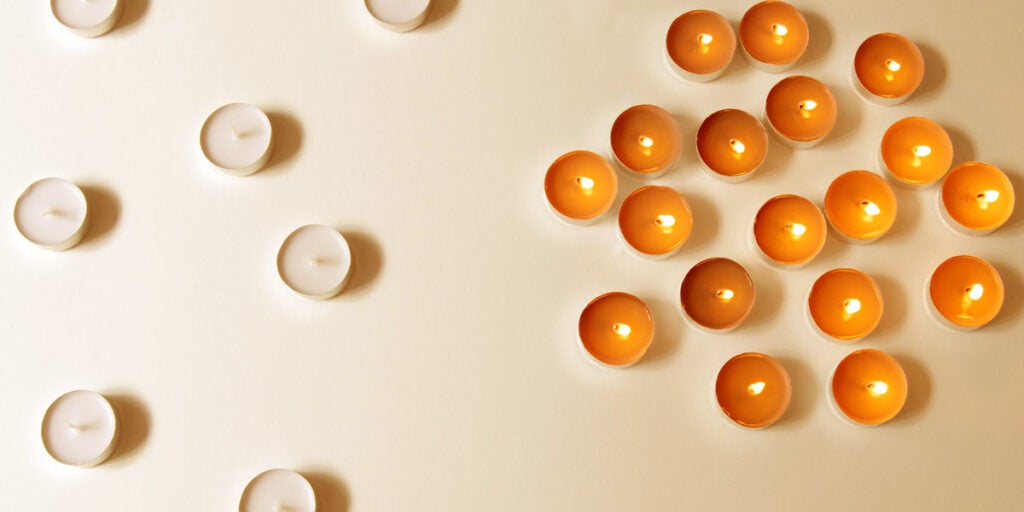 Seven unlit, white votive candles are separated on one side of surface. On the other, a cluster of lit, orange votive candles huddle together.