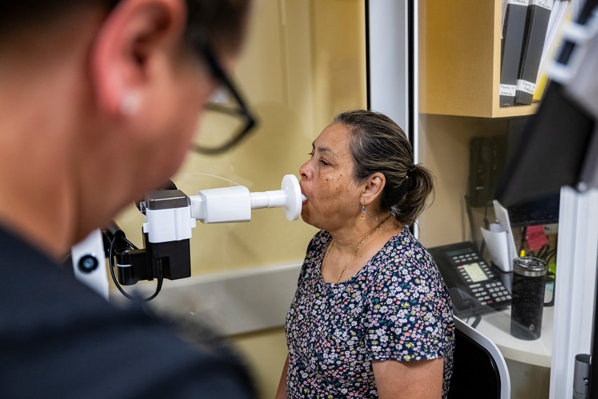 A middle-aged Latina patient sits in a booth and breathes into a spriometer to test her lung function at a medical clinic. A technician monitors the process.