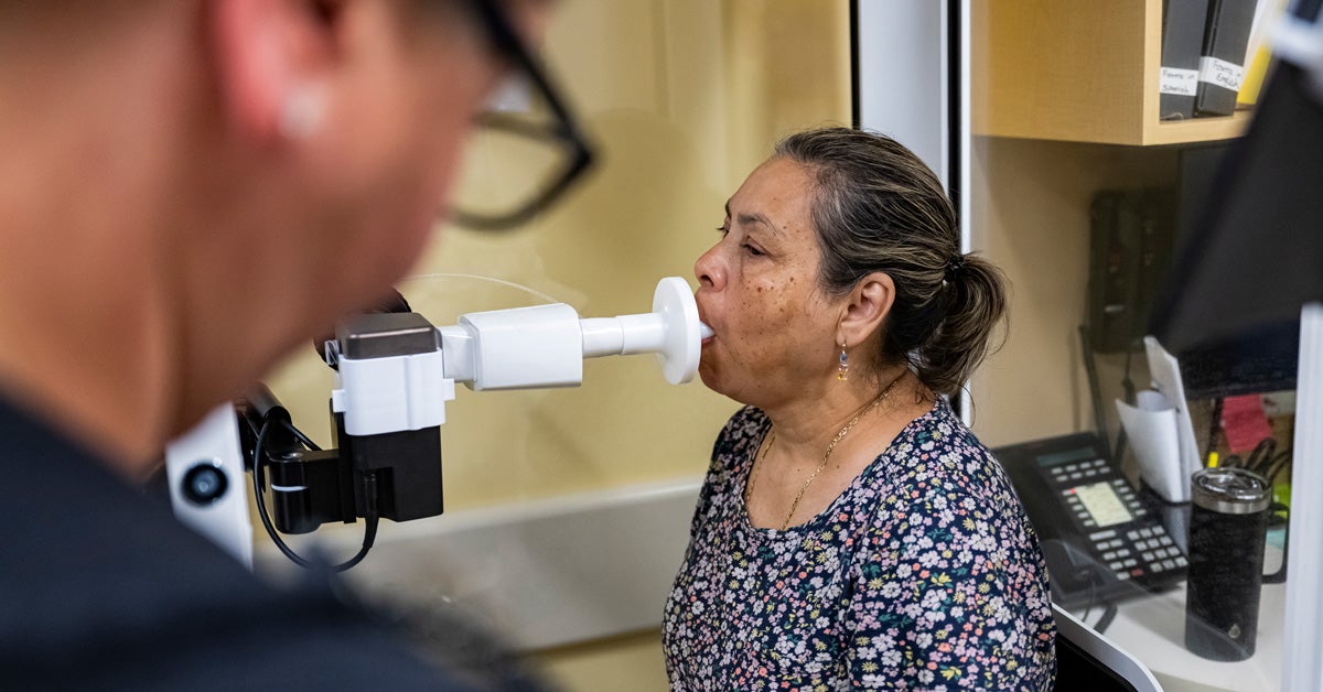 A middle-aged Latina patient sits in a booth and breathes into a spriometer to test her lung function at a medical clinic. A technician monitors the process.