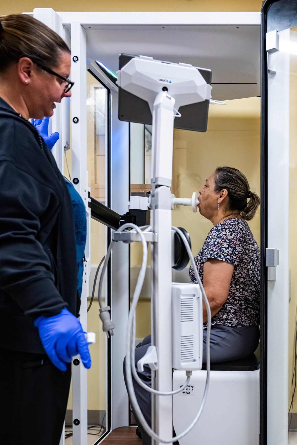 A middle-aged Latina patient sits in a booth and breathes into a spriometer to test her lung function at a medical clinic. A technician stands beside, monitoring the process.