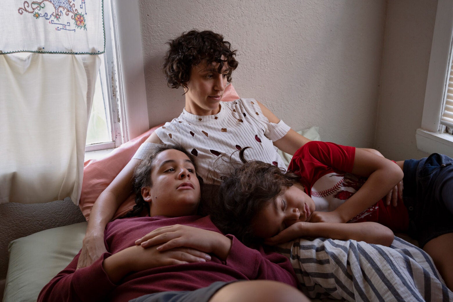 A middle aged mother reclines on a bed with her two male children resting on her legs and nearby. Light shines through a window by her shoulder.