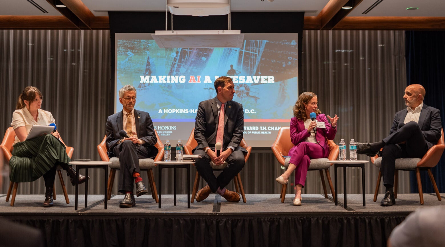 Five panelists sit on a stage in conversation. Behind them is a screen that reads "Making AI a Lifesaver: A Hopkins-Harvard event in D.C."