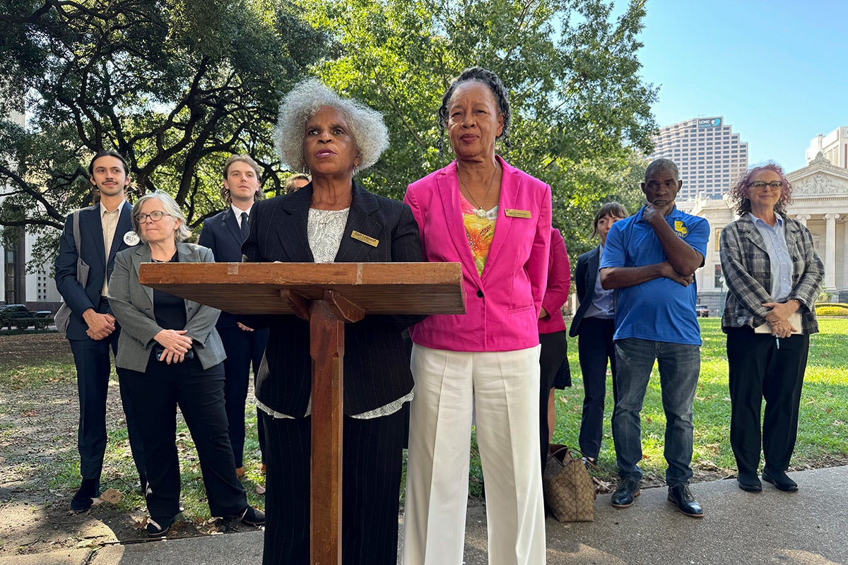 Gail LeBoeuf and Barbara Washington stand at a podium. New Orleans buildings and a large tree are visible in the background.