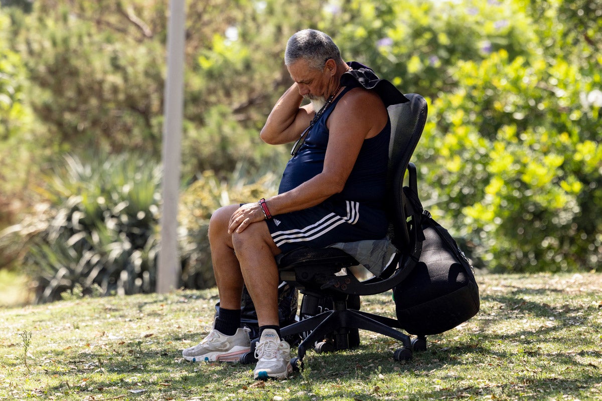 A man sitting in a black office chair in a shady park wipes his neck. He wears shorts, sneakers and a tank top.